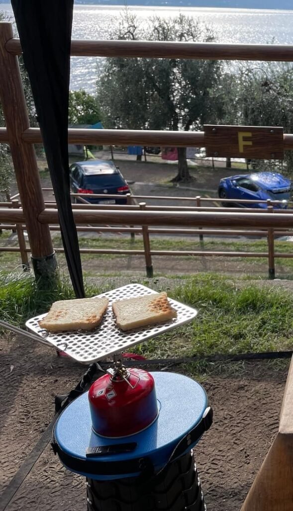 Soto-Campingkocher mit Toasteraufsatz und zwei Toasts steht direkt auf dem Have a Seat Hocker – Frühstücksszene auf einem Campingplatz mit Blick auf den Gardasee.