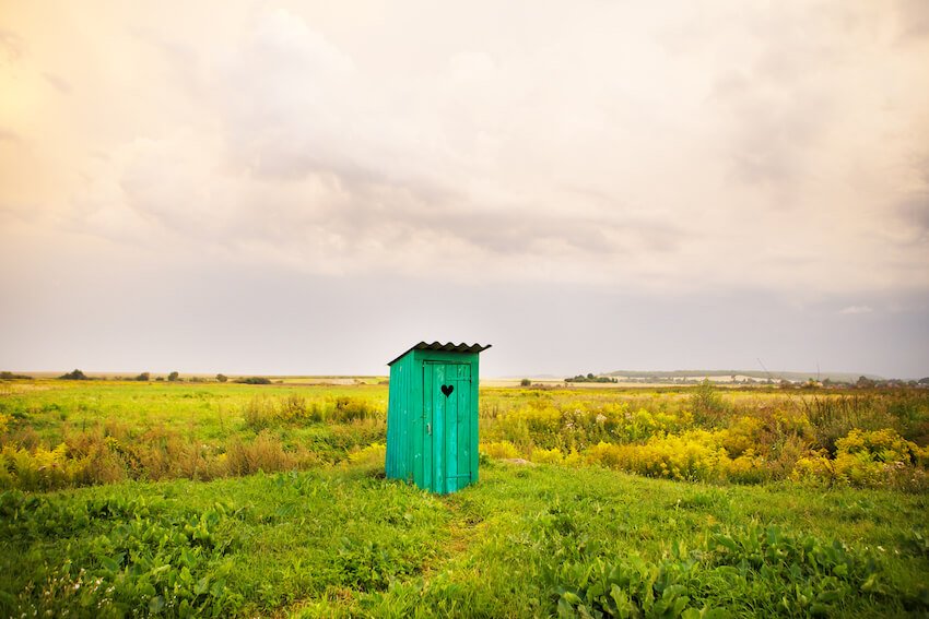 Holztoilettenhäuschen mit Herz in der Tür auf grüner Wiese unter bewölktem Himmel – Symbolbild für Campingtoiletten