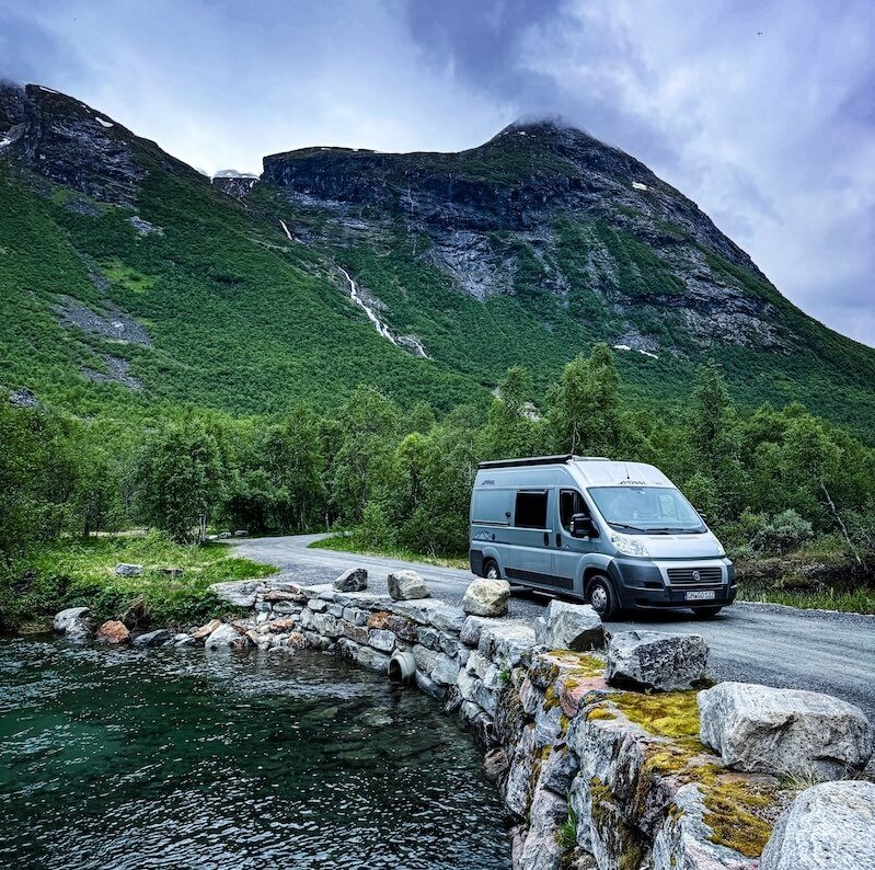 Pössl Camper auf einer kleinen Brücke über klarem Wasser, im Hintergrund hohe grüne Berge unter bewölktem Himmel.