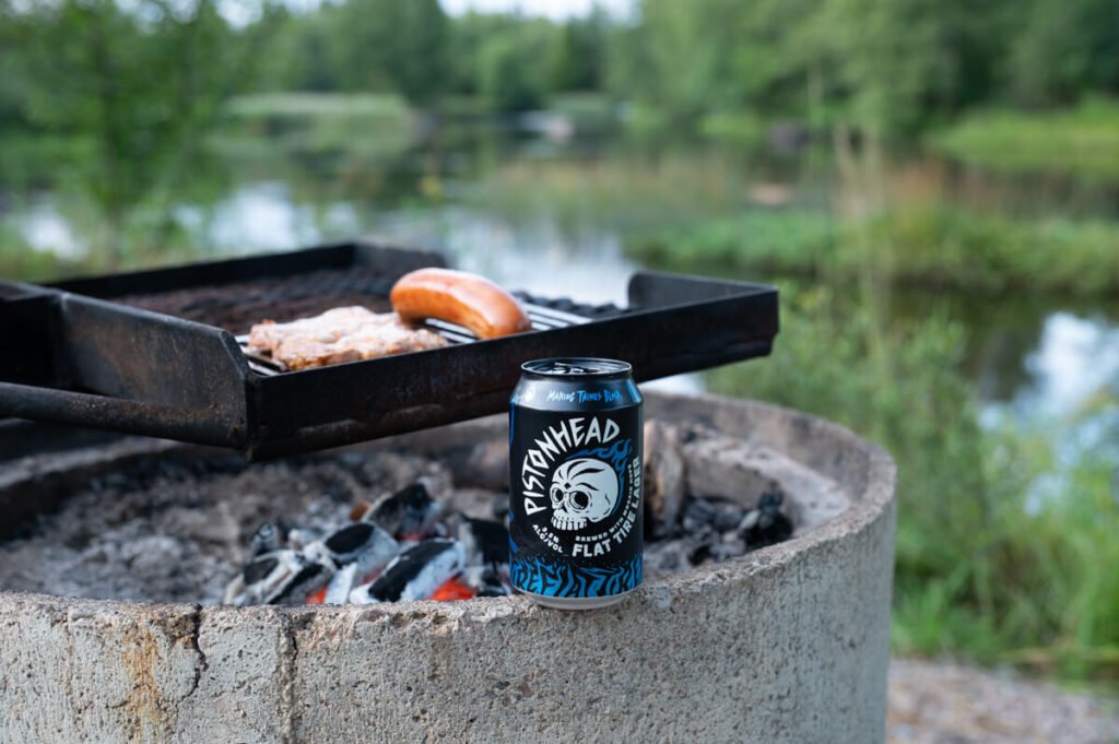 Grillstelle aus Beton mit Fleisch und Wurst über offenem Feuer, im Vordergrund eine Dose Pistonhead Flat Tire Lager – einsamer Stellplatz am Fluss in Schweden.