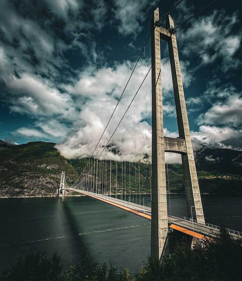 Imposante Aufnahme der Hardangerbrücke in Norwegen mit dramatischem Himmel und Fjordlandschaft im Hintergrund.