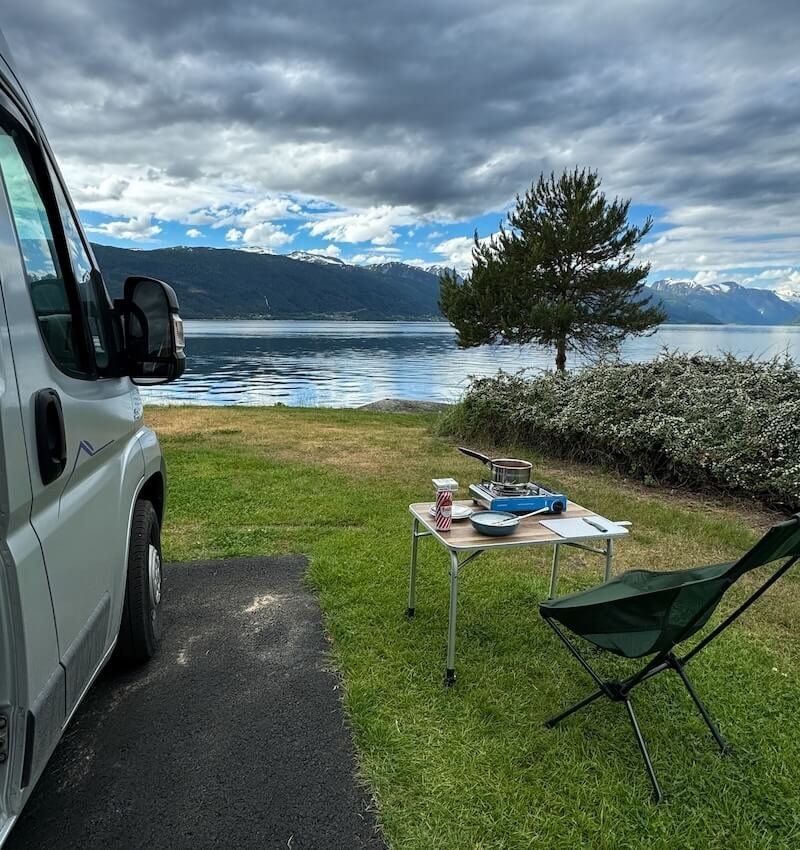 Stellplatz direkt am norwegischen Fjord mit Blick auf schneebedeckte Berge. Campingtisch mit Gaskocher, Topf, Teller und Dose Früh Kölsch. Links im Bild der Camper, rechts ein Helinox-Stuhl. Perfekte Outdoor-Küche mit Panorama.
