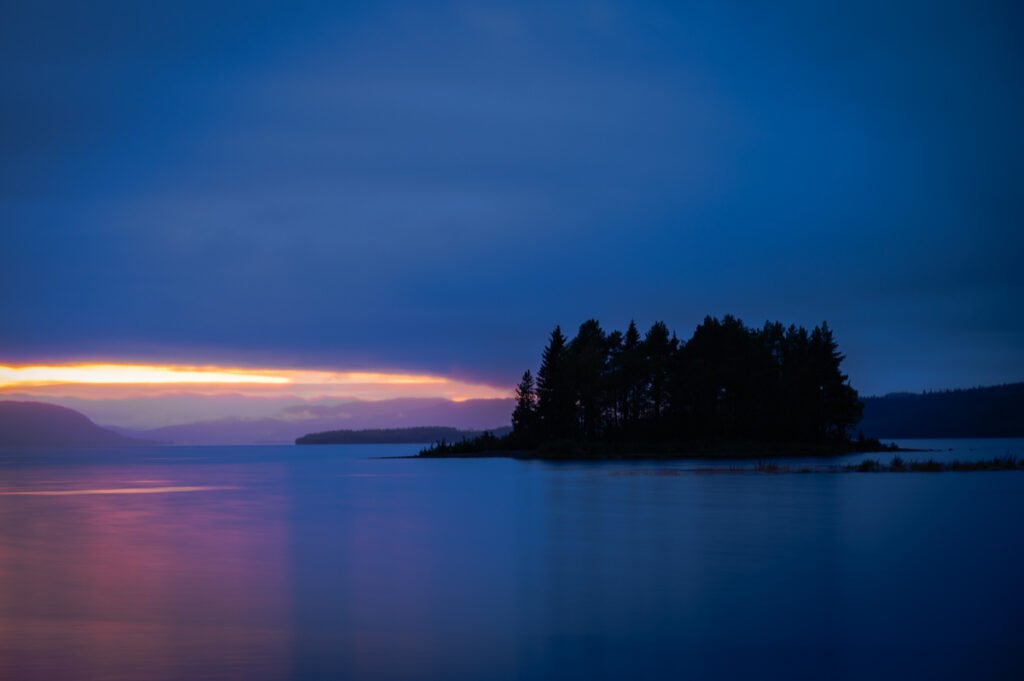 Langzeitbelichtetes Foto eines Sees bei Dämmerung mit Bauminsel und dramatischem Himmel in Skandinavien.