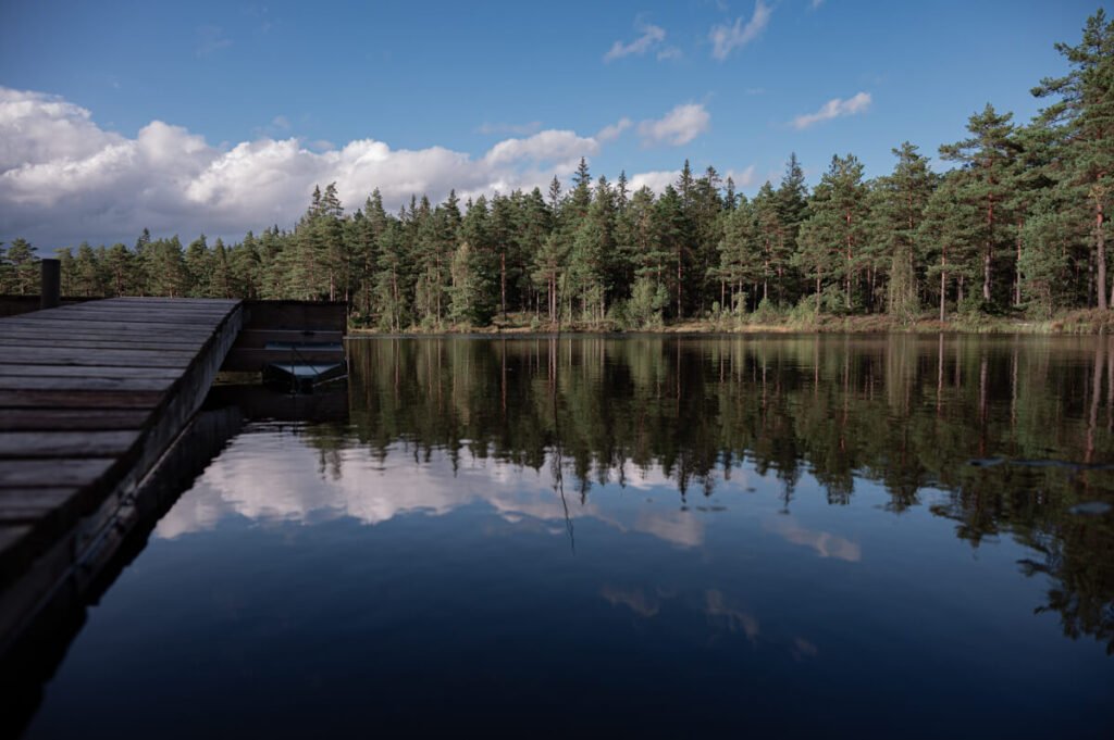 Holzsteg an einem ruhigen See in Schweden, umgeben von Wald, mit Spiegelung der Wolken im Wasser