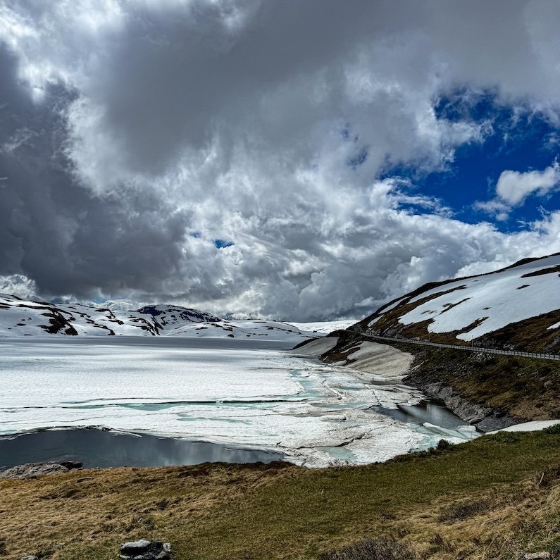 Zugefrorener Bergsee mit Schneefeldern und dramatischer Wolkenkulisse auf einer Passstraße nahe Vik in Norwegen