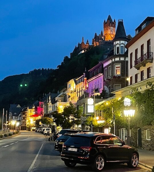 Abendliche Straßenansicht in Cochem mit beleuchteter Moselpromenade und der Reichsburg auf dem Hügel im Hintergrund.