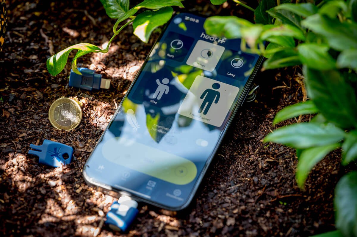 Heat it Insektenstichheiler am iPhone im Einsatz mit App, USB-C und Lightning Version im Größenvergleich mit 1-Euro-Münze